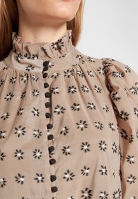 Beige paita, jossa on röyhelökaulus, koristeena musta kukkakirjonta ja mustat napit edessä. Teksturoitu kangas ja puffi hihat.