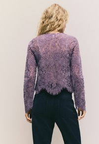 Lila Spitzenoberteil mit langen Ärmeln, gewelltem Saum, floralen Mustern, transparenter Textur, getragen über dunkler Jeans, Rückansicht.