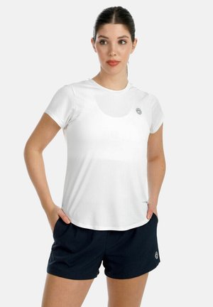 Femme portant un t-shirt de sport blanc à manches courtes et un short noir, debout les mains dans les poches, regardant légèrement sur le côté.