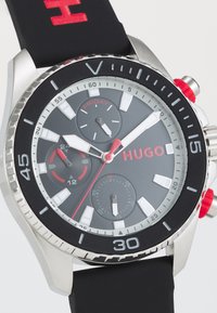 Black rubber strap watch with stainless steel case. Grey dial features red accents, three subdials, and a date display. Bold hour markers.