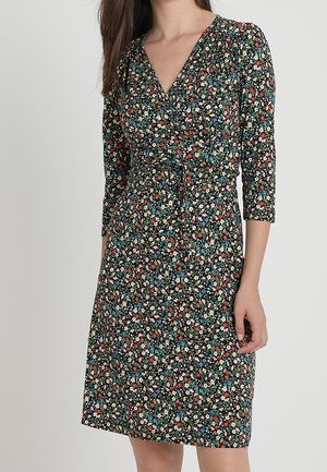 Femme portant une robe imprimée florale jusqu'aux genoux, manches trois-quarts et col en V, debout devant un fond clair uni.