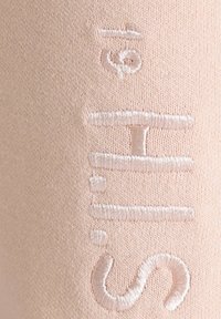 Hellrosa Textil mit dezent gesticktem, weißem Text und einzigartigen Zeichen. Glatte Textur mit einem weichen, dehnbaren Erscheinungsbild.