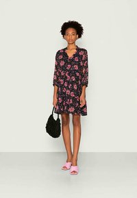 Vestido floral com base preta, flores rosas; corpete justo, saia rodada, mangas três quartos. Combinado com uma mala preta texturizada e sandálias rosas.