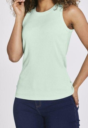 Kvinde iført lysegrøn ærmeløs ribbet top og mørkeblå jeans, med hånden hvilende nær ansigtet og på hoften.