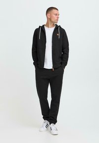 Mann steht mit Händen in den Taschen, trägt einen schwarzen Zip-Hoodie, schwarze Hose, ein weißes T-Shirt und weiße Sneakers mit schwarzen Streifen, schaut nach rechts.