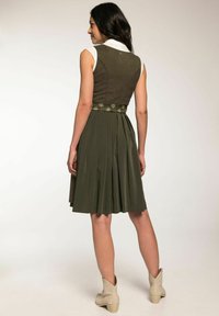 Robe sans manches verte avec un buste ajusté, jupe plissée et une ceinture fleurie. Accentuée d'un col blanc et de bottines beiges.