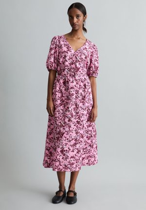 Femme portant une robe midi rose à fleurs avec des manches bouffantes et des chaussures Mary Jane noires, debout devant un fond gris uni.