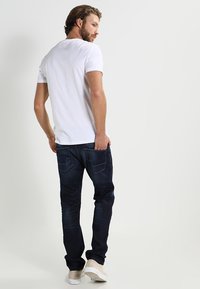Männliches weißes Baumwoll-T-Shirt kombiniert mit dunkelblauen Jeans. Die Jeans haben Gesäßtaschen mit Akzentnähten und eine taillierte Passform.