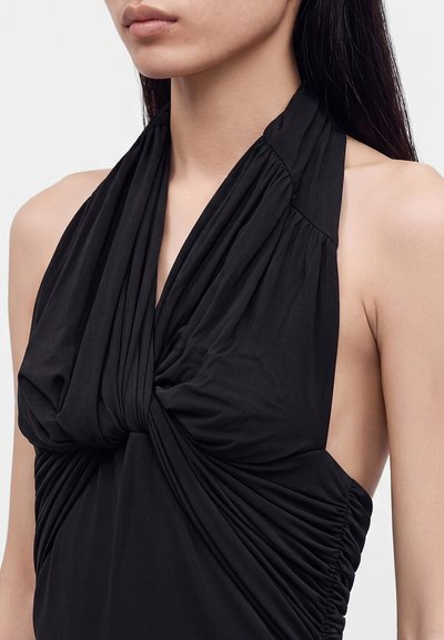 Γυναίκα που φοράει μαύρο αμάνικο halter top με στριφτό και ρυτιδιασμένο ύφασμα μπροστά και στα πλάγια, με φόντο μονόχρωμο.