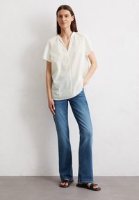 Kurzärmlige, kragenlose weiße Bluse, kombiniert mit blauen Schlagjeans. Weicher Stoff mit lockerem Schnitt; entspannte Silhouette. Schwarze Sandalen runden das Outfit ab.