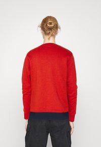 Sudadera roja de manga larga, con dobladillo y puños acanalados; presenta una textura suave y una banda inferior de color azul marino en contraste.