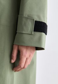 Tessuto del cappotto verde con una striscia di Velcro nera testurizzata al polsino. Il polsino presenta una toppa rettangolare e un taglio pulito.