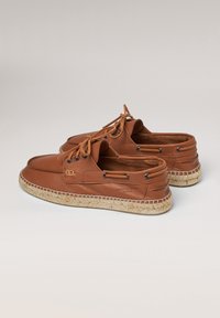 Zapatos de barco de cuero marrón con un acabado texturizado, suelas de goma y detalles de yute trenzado alrededor del borde. Presenta ojetes y cordones negros.