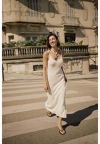 Abito maxi bianco, lavorato a maglia, senza maniche, con un motivo testurizzato; indossato con infradito nere; mentre cammina su un attraversamento pedonale con uno sfondo chiaro.
