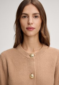Giovane donna con lunghi capelli castani che indossa un cardigan a maglia beige con due bottoni dorati decorativi, fronteggiando l'obiettivo con un'espressione neutra.