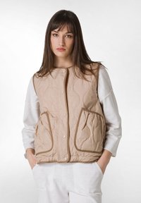 Beige gesteppte Weste mit zwei vorderen Taschen, Druckknopfverschluss und kontrastierendem Besatz. Getragen über einem weißen Langarmoberteil.
