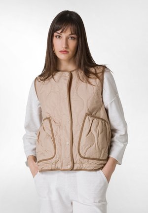 Beige liivi, jossa on painettu kuosi, kaksi etutaskua, nappisuljin ja erottuva reunus. Päällä valkoinen pitkähihainen paita.