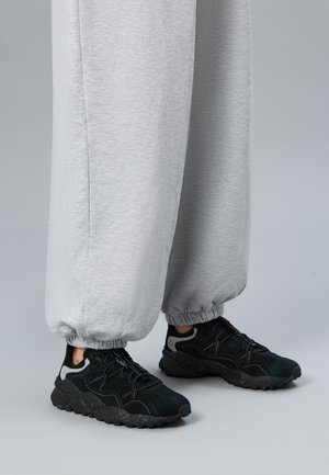 Legs wearing light gray sweatpants and black athletic shoes with textured soles, standing on a gray surface against a plain background.