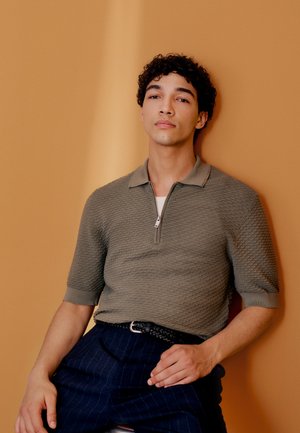 Joven con cabello rizado que lleva una camisa polo verde oliva texturizada y pantalones oscuros a rayas, sentado frente a un fondo beige.