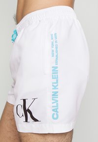 Shorts de bain blancs avec une taille élastique, cordon bleu, grand logo "CK" noir et texte "CALVIN KLEIN" en bleu sur le côté.