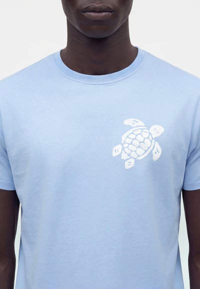 Homme portant un t-shirt bleu clair avec un motif de tortue de mer blanche brodée sur la poitrine gauche, visible du cou jusqu'au milieu du torse.