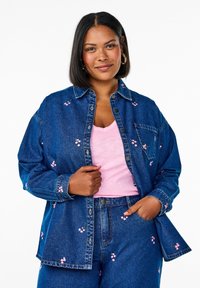 Chaqueta de mezclilla con bordado floral rosa, cierre de botones en la parte delantera, dos bolsillos en el pecho, combinada con una camiseta de pico rosa claro y pantalones de mezclilla.