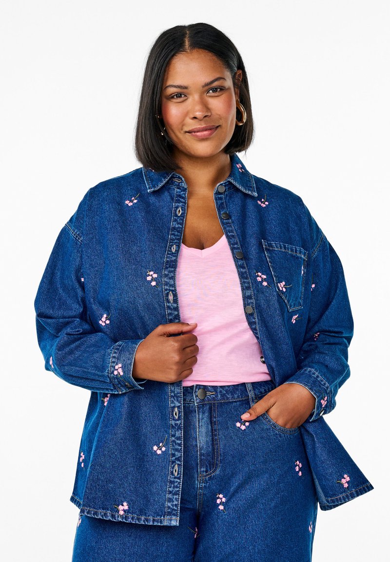 Chaqueta de mezclilla con bordado floral rosa, cierre de botones en la parte delantera, dos bolsillos en el pecho, combinada con una camiseta de pico rosa claro y pantalones de mezclilla.