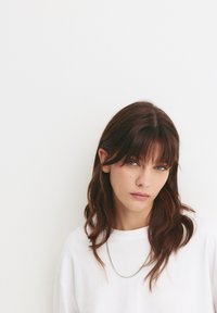 Weißes, übergroßes T-Shirt, wellige, lange braune Haare mit Pony, feine Silberkette, direkter Blick, gedämpfter Hintergrund.