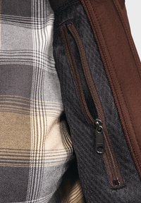 Das Innere der braunen und schwarzen Reißverschlussjacke verfügt über ein graues und tanfarbendes Karolining. Der strukturierte Stoff steht im Kontrast zu den glatten Abschnitten.