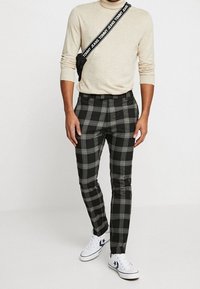 Muž v béžovém svetru, černobíle kostičkovaných kalhotách, bílých teniskách s černou hvězdou a černé kabelce přes rameno se popruhem "Tommy Jeans".