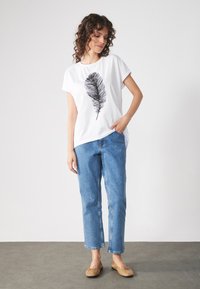 Hvit t-skjorte med svart fjærtrykk, satt sammen med lyseblå rette jeans og brune leiligheter, mot en ensfarget bakgrunn.