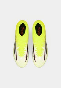 Λαμπερά κίτρινα ποδοσφαιρικά παπούτσια με μαύρες και κόκκινες λεπτομέρειες, υφασμάτινη επάνω επιφάνεια, λεπτός σχεδιασμός και επίπεδα κορδόνια. Διαθέτουν χαρακτηριστικό γεωμετρικό μοτίβο.