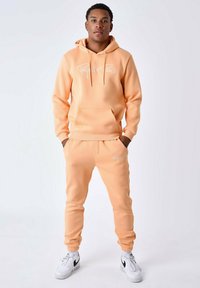Sudadera y pantalones de chándal en color melocotón, confeccionados en tejido suave, que incluyen un bolsillo frontal y un logo en blanco. El calzado es blanco con detalles en negro.
