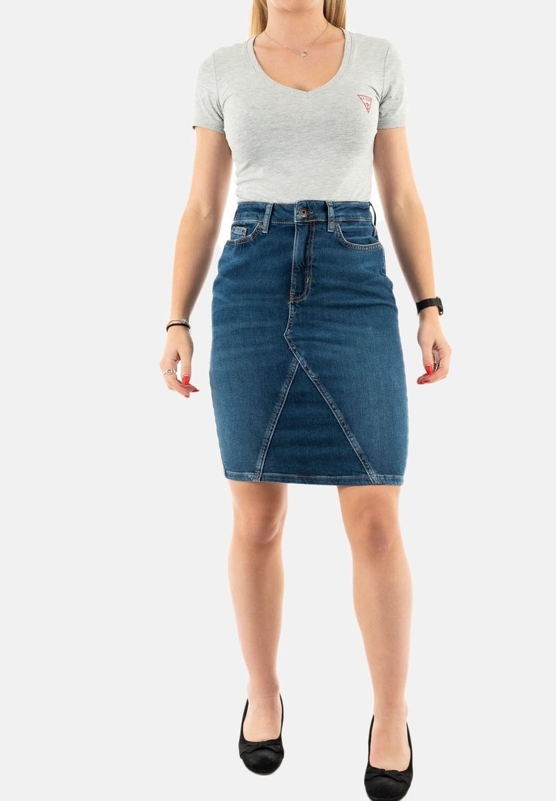 Femme portant un t-shirt gris clair à col en V et une jupe en jean bleu taille haute avec un détail de couture à l'avant, associée à des ballerines noires.