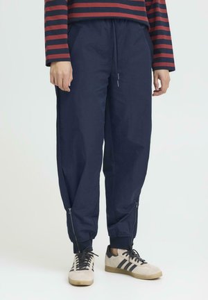 Navy-blue tapered pants made from lightweight fabric, featuring zippered cuffs and side pockets, paired with light-colored shoes.