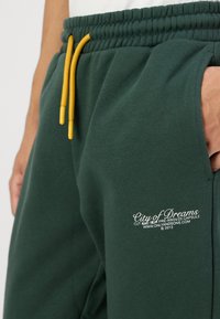 Green cotton sweatpants with side pockets, elastic waistband, and yellow drawstrings. Features white embroidered text on the left thigh.