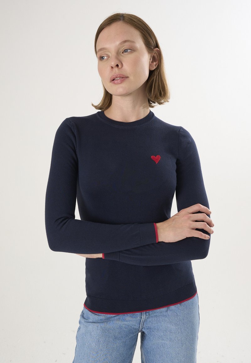 Marineblauer Langarmpullover mit rotem Herzstickerei auf der linken Brust, mit roten Akzenten an den Bündchen und am Saum. Weiche Textur.