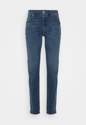 Jean slim bleu avec fermeture avant par bouton et zip, passants de ceinture et design classique à cinq poches sur un fond clair uni.