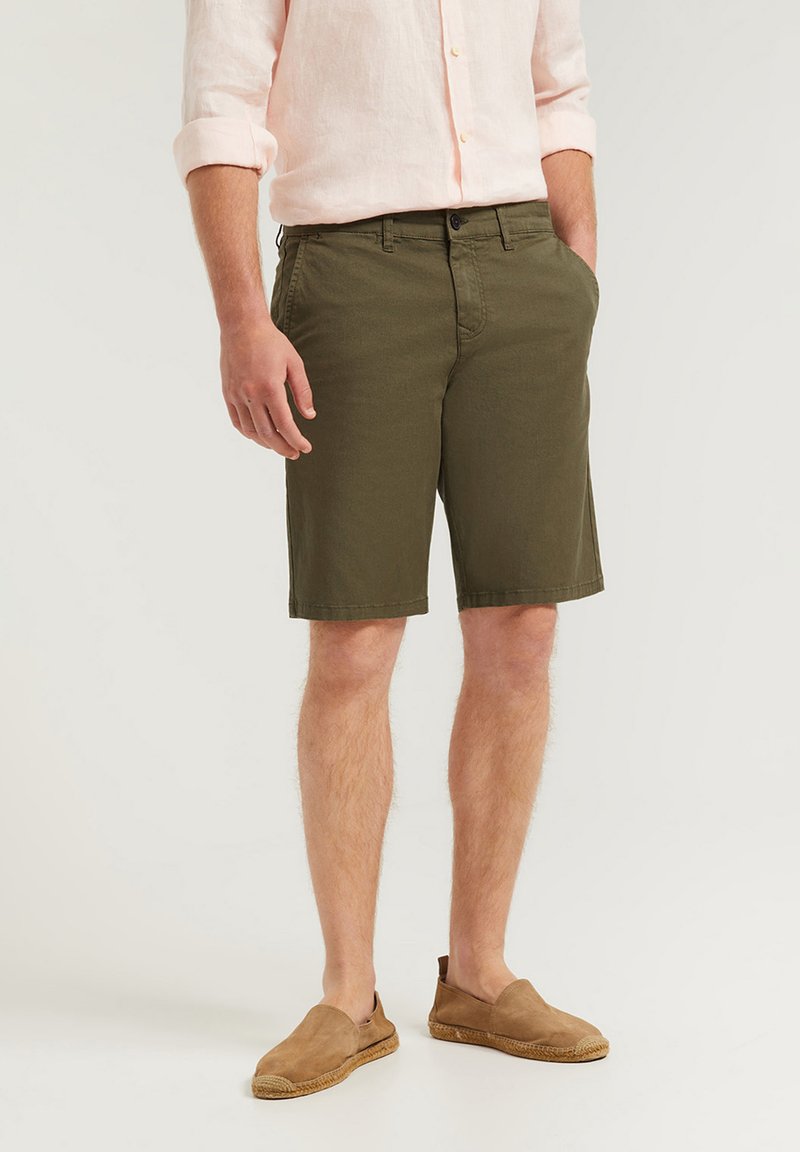 Olivgröna bomullsshorts med rak passform, utrustade med sidofickor, knappstängning och fållade kanter. Bärs med beiga slip-on skor.