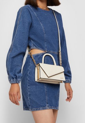 Vrouw draagt een blauwe denim jurk met lange mouwen en een uitgesneden taille, draagt een kleine crèmekleurige schoudertas met gouden kettingaccenten.