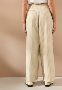 Beige weite Hosen mit vertikalen Streifen. Besitzt einen hohen Bund, Seitentaschen und einen strukturierten Stoff. Stilisiert mit cremefarbenen Schuhen.