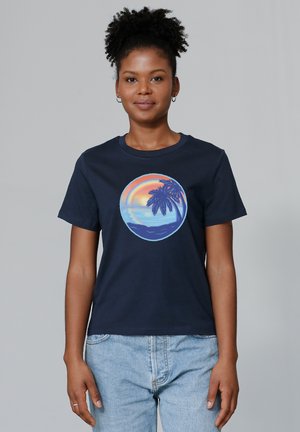 T-shirt en coton bleu marine avec un graphique circulaire représentant un coucher de soleil avec des palmiers, un océan et un ciel coloré. Manches courtes et col rond.