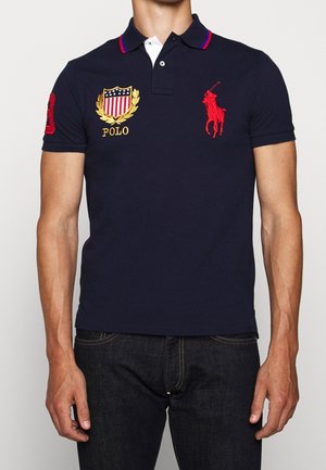 Dunkelblaues Polohemd aus Baumwolle mit weißem Kragen, roten Akzenten, gesticktem Logo und Aufnähern an den Ärmeln. Standardpassform.