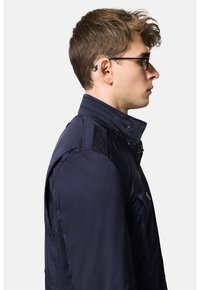 Granatowa ocieplana kurtka z wysokim kołnierzem, zamek błyskawiczny i zatrzaskami, oraz bocznymi kieszeniami. Posiada gładką, błyszczącą fakturę i dopasowany krój.