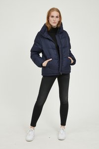 Veste rembourrée bleu marine avec un col montant, une fermeture éclair et des poches latérales. Présente un motif matelassé et est portée par-dessus un haut noir et un jean.
