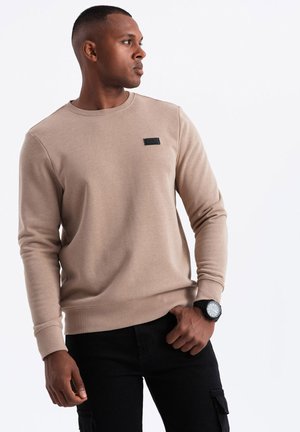 Beige huppari, jossa on pyöreä kaula-aukko ja pieni musta etiketti rinnassa. Varustettu resorilla varustetuilla hihansuilla ja helmalla. Yhdistetty mustiin cargo-housuihin.
