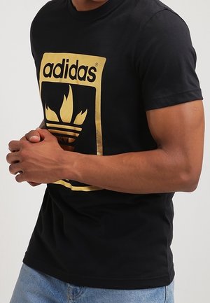 Mann trägt ein schwarzes Adidas-T-Shirt mit goldenem Logo und blaue Jeans, die Hände vorne gefaltet.
