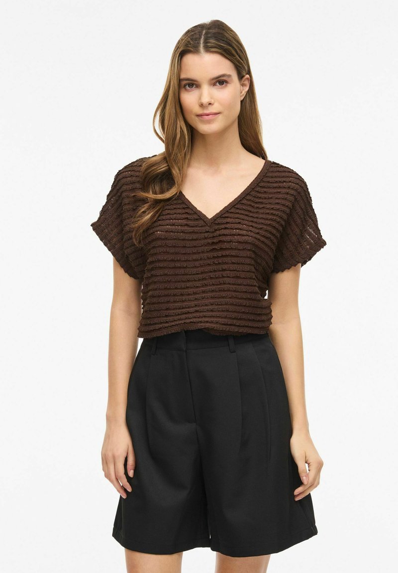 Jeune femme aux longs cheveux bruns portant un haut marron texturé à col en V et manches courtes, ainsi qu'un short noir taille haute longueur genou.