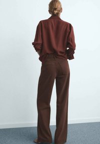 Chemise marron à manches longues avec des poignets retroussés, assortie à un pantalon large à taille haute marron. Tissu lisse avec poches arrière et design minimaliste.