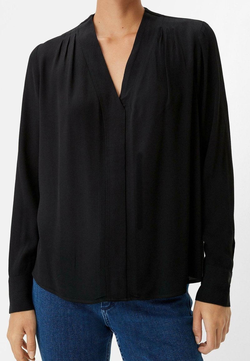 Blouse noire à manches longues avec plis près des coutures des épaules, encolure en V, portée avec un jean bleu, modèle cadré au niveau du cou et des poignets.
